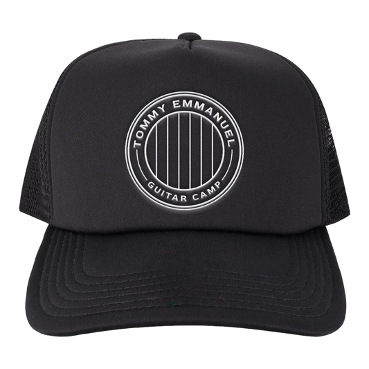 A black trucker hat with a white circular embroidered logo on the front that reads 'TOMMY EMMANUEL GUITAR CAMP'. The hat features a structured black front panel, a curved black brim, and black mesh back panels, shown against a plain white background.